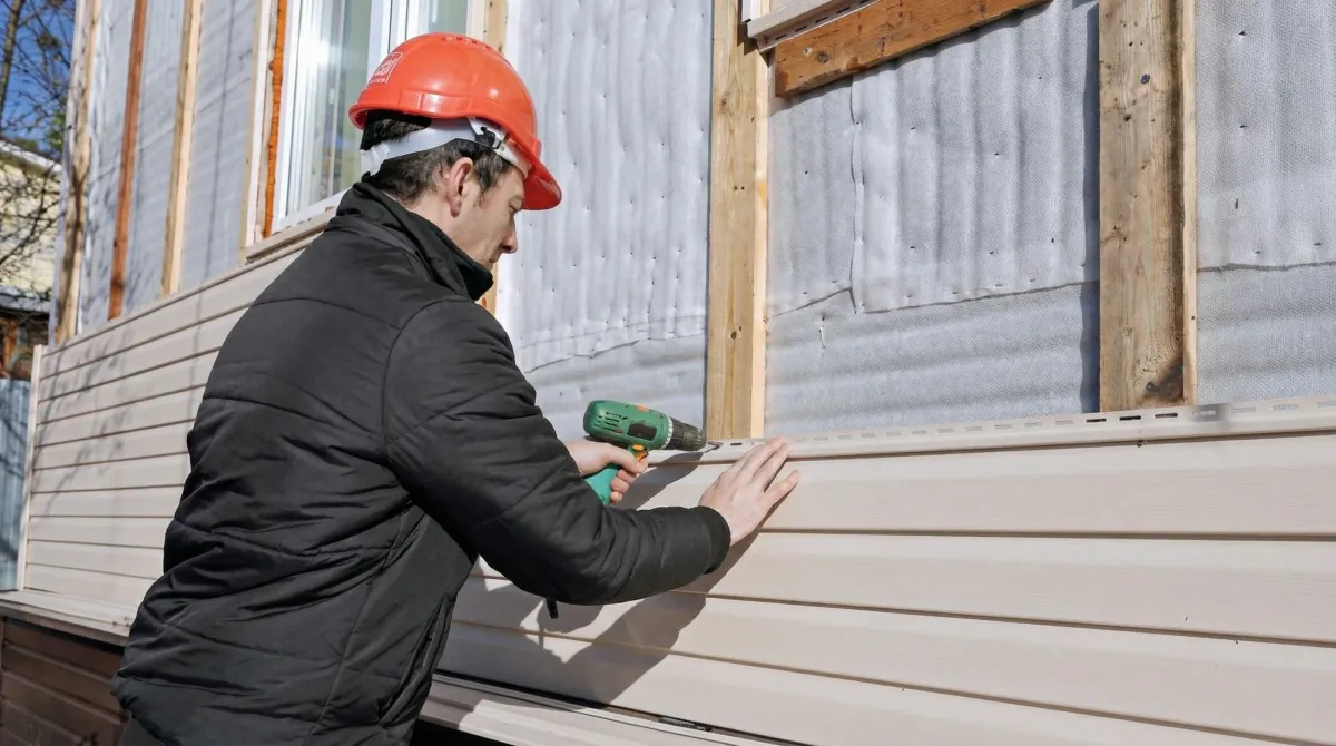 Siding replacement professional using a power drill to install new tan vinyl siding on a home exterior in Houston, TX